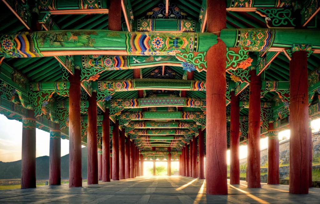 Jinnamgwan. Interior de un corredor tradicional coreano con columnas de madera roja y vigas decoradas con dancheong, el estilo policromado característico de la arquitectura de Joseon. La luz entra lateralmente y resalta los patrones geométricos y florales del techo, mostrando la estructura interna típica de los grandes pabellones administrativos del periodo.